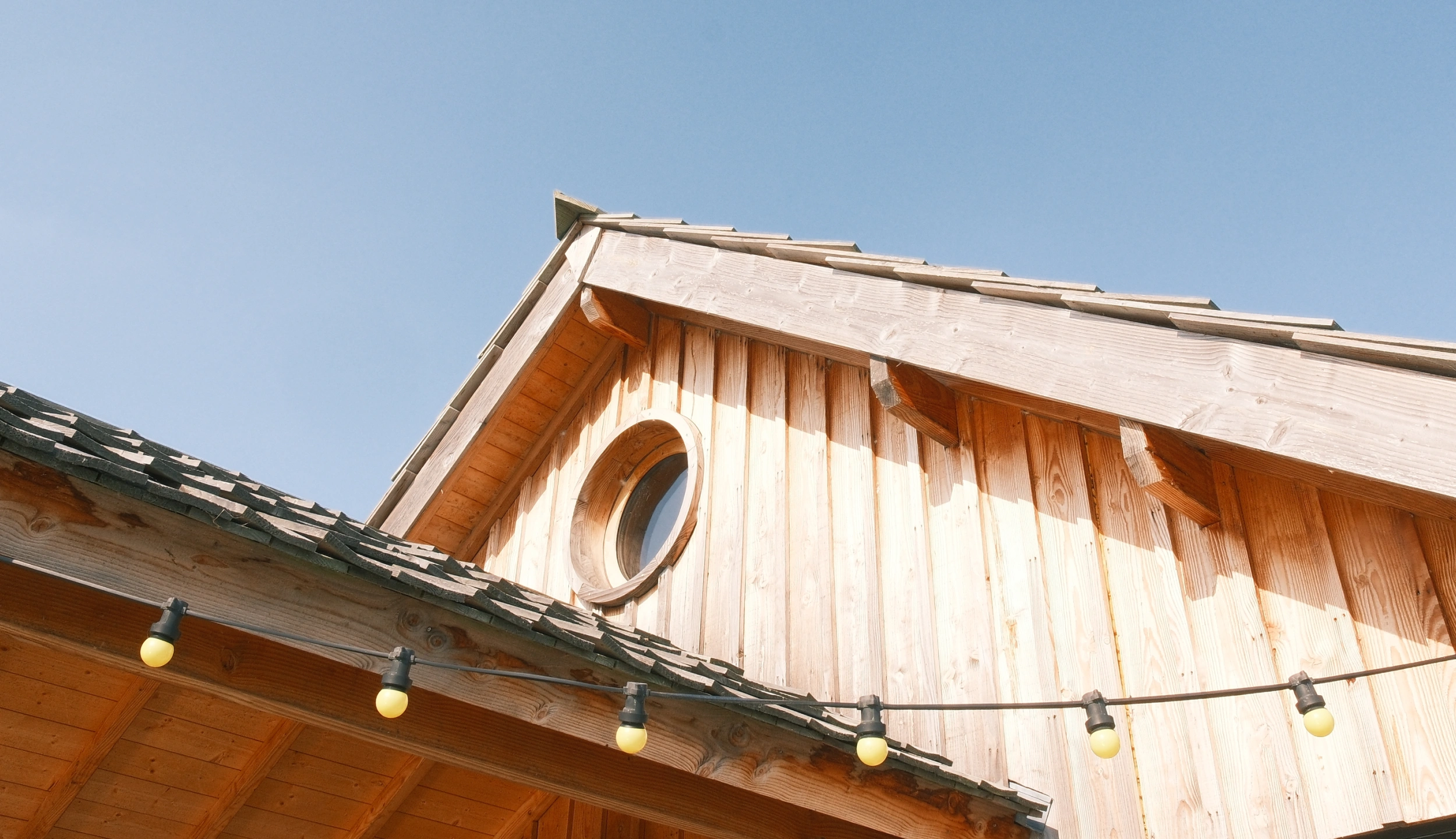 Pignon en bois clair d'une cabane avec fenêtre ronde et guirlande lumineuse, sous un ciel bleu